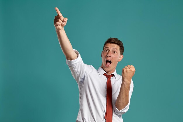 Retrato de un hombre moreno feliz con ojos marrones, vestido con una clásica camisa blanca y corbata roja. Está actuando como si hubiera ganado algo posando en un estudio sobre un fondo azul. Concepto de gesticulación