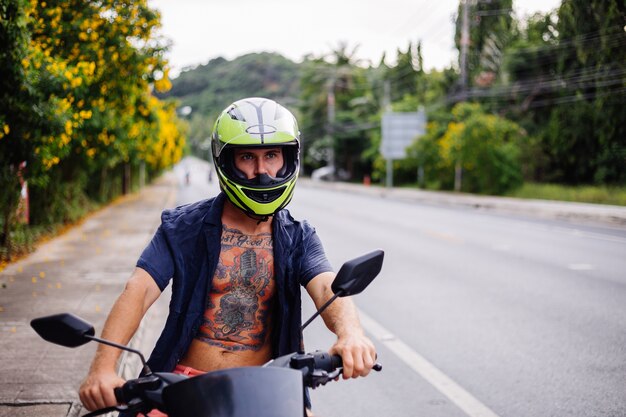 Retrato de hombre motorista tatuado en casco amarillo en moto en el lado de una carretera muy transitada en Tailandia