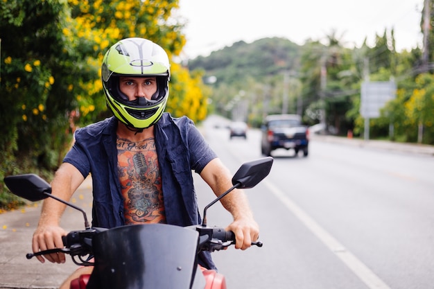 Retrato de hombre motorista tatuado en casco amarillo en moto en el lado de una carretera muy transitada en Tailandia