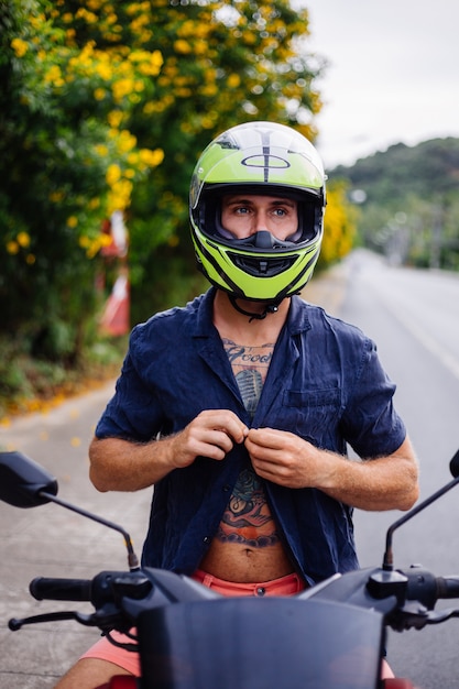 Retrato de hombre motorista tatuado en casco amarillo en moto en el lado de una carretera muy transitada en Tailandia