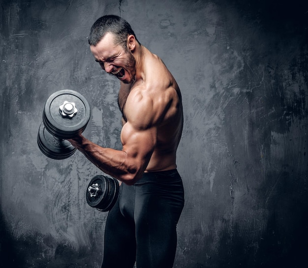 Retrato de hombre musculoso sin camisa haciendo ejercicios de bíceps sobre fondo gris.