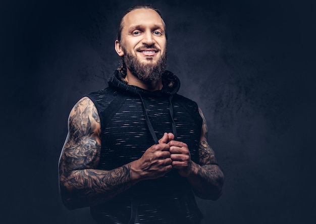 Retrato de un hombre musculoso tatuado con barba y un elegante corte de pelo con ropa deportiva negra. Aislado en un fondo oscuro.