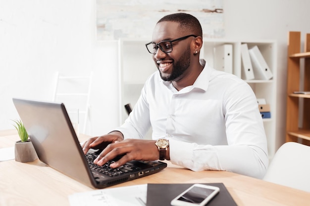 Retrato de un hombre de negocios afroamericano sonriente con camisa blanca y anteojos sentado y trabajando en su computadora portátil en la oficina aislado