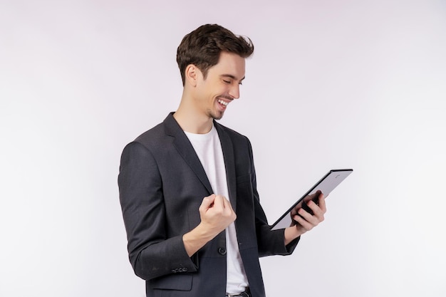 Retrato de un hombre de negocios alegre y atractivo haciendo un gesto de ganador apretando el puño y usando una aplicación de dispositivo buscando en la web aislada sobre fondo de color blanco