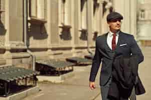 Foto gratuita retrato de un hombre de negocios árabe inglés retro de la década de 1920 con traje oscuro, corbata y gorra plana caminando por calles antiguas