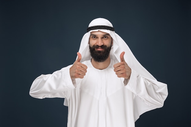 Retrato de hombre de negocios de Arabia Saudita. Modelo masculino joven de pie mostrando un gesto de pulgar hacia arriba. Concepto de negocio, finanzas, expresión facial, emociones humanas.