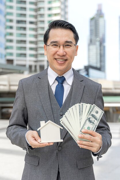 Retrato de hombre de negocios asiático con dinero billetes de un dólar estadounidense y casa modelo en el distrito de negocios