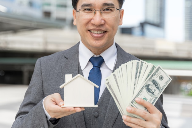 Retrato de hombre de negocios asiático con dinero billetes de un dólar estadounidense y casa modelo en el distrito de negocios