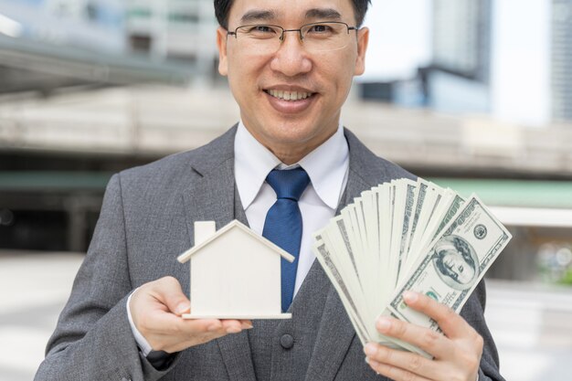 Retrato de hombre de negocios asiático con dinero billetes de un dólar estadounidense y casa modelo en el distrito de negocios
