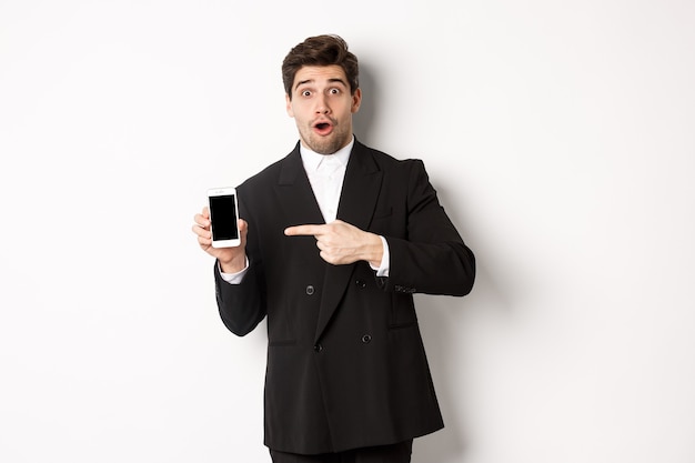 Retrato de hombre de negocios atractivo en traje negro, mirando sorprendido y señalando con el dedo a la pantalla del teléfono inteligente, de pie sobre fondo blanco.