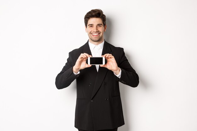 Retrato de hombre de negocios atractivo en traje negro, sosteniendo el teléfono inteligente horizontalmente y mostrando la pantalla, sonriendo complacido, de pie contra el fondo blanco.