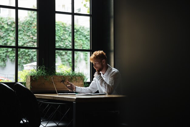 Retrato de hombre de negocios barbudo pelirrojo serio con smartphone, sentado en el lugar de trabajo