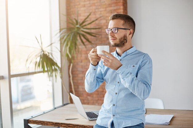 Retrato de hombre de negocios caucásico sin afeitar maduro en gafas y camisa clásica de pie en la oficina de luz, beber café, relajarse durante las vacaciones. Concepto de negocio.