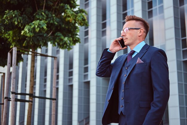 Retrato de un hombre de negocios elegante y confiado vestido con un elegante traje habla por teléfono y mira hacia otro lado mientras está de pie al aire libre contra un fondo de rascacielos.