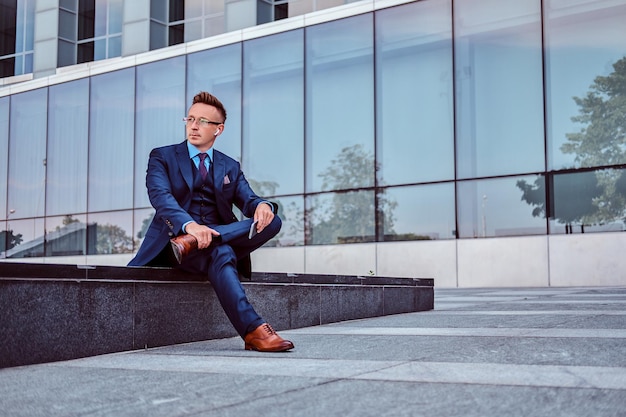 Retrato de un hombre de negocios elegante y confiado vestido con un traje elegante sostiene un teléfono inteligente y mira hacia otro lado mientras se sienta al aire libre contra el fondo de un rascacielos.