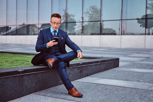 Retrato de un hombre de negocios elegante y confiado vestido con un traje elegante usando un teléfono inteligente mientras se sienta al aire libre contra un fondo de rascacielos.