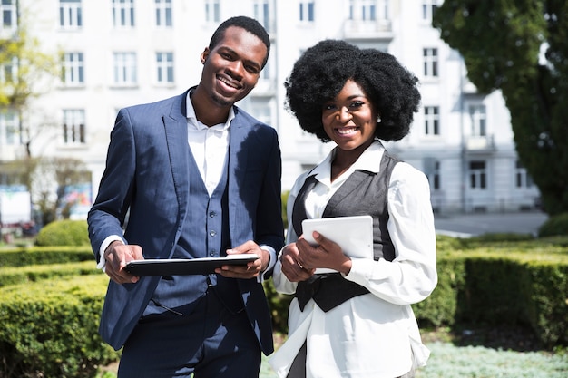 Retrato de un hombre de negocios y de una empresaria jovenes africanos que sostienen el tablero y la tableta digital que miran la cámara