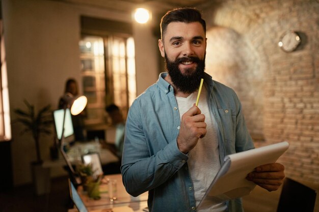 Retrato de hombre de negocios feliz analizando informes mientras trabaja hasta tarde en la oficina