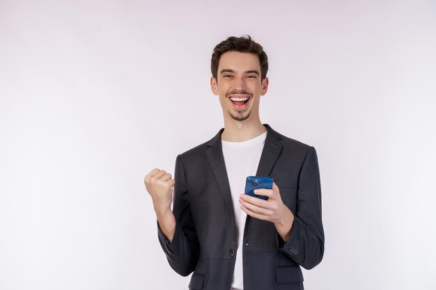 Retrato de un hombre de negocios feliz que usa un teléfono inteligente y hace un gesto de ganador apretando el puño sobre fondo blanco