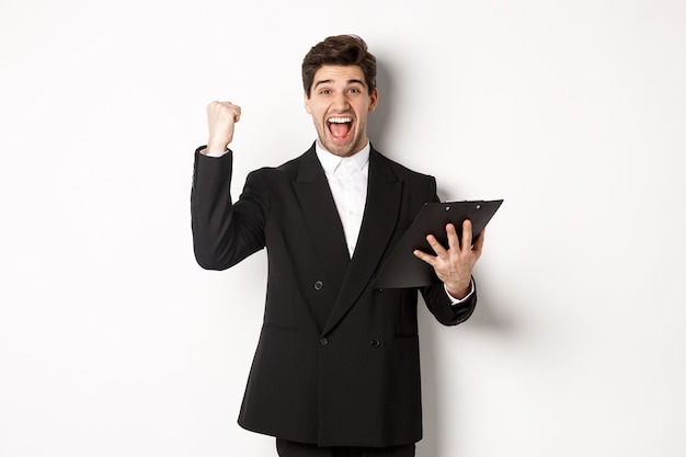 Retrato de hombre de negocios guapo emocionado en traje negro, sosteniendo el portapapeles y haciendo bomba de puño, lograr la meta y regocijarse, de pie sobre fondo blanco.