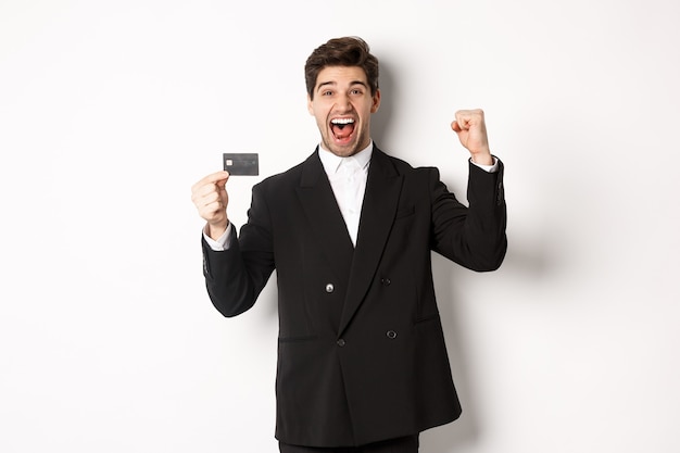 Retrato de hombre de negocios guapo emocionado en traje, regocijo y mostrando tarjeta de crédito, de pie contra el fondo blanco.