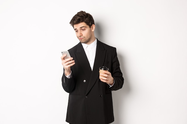 Retrato de hombre de negocios guapo pensativo, tomando café y navegando en internet, mirando la pantalla del teléfono inteligente, de pie contra el fondo blanco.