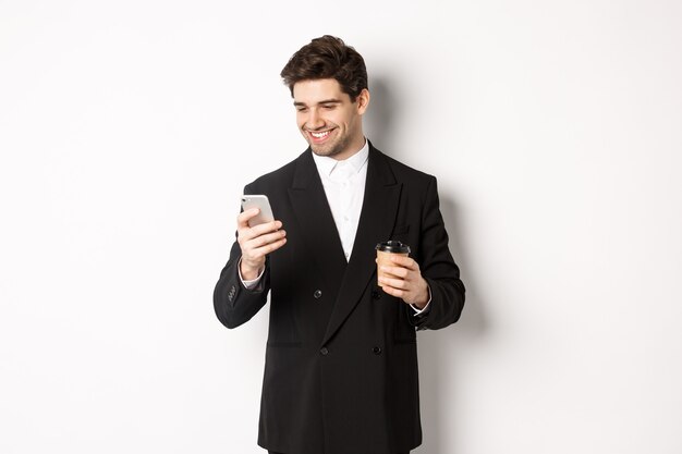 Retrato de hombre de negocios guapo y seguro en traje negro, tomando café y usando el teléfono móvil, sonriendo complacido, de pie sobre fondo blanco.