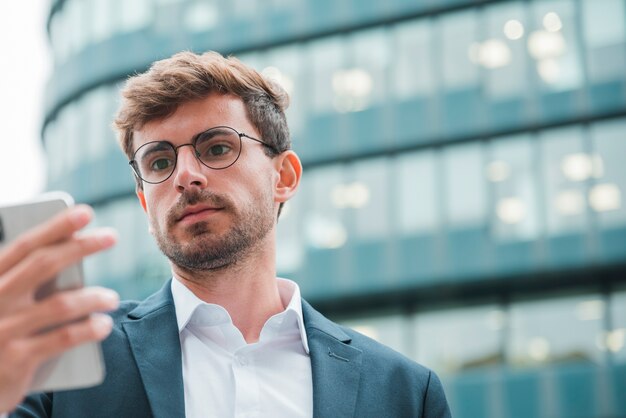 Retrato de un hombre de negocios joven que mira el teléfono móvil que se coloca delante del edificio corporativo