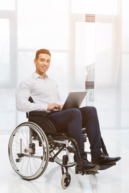 Retrato de un hombre de negocios joven sonriente discapacitado que se sienta en la silla de ruedas usando el ordenador portátil en la oficina