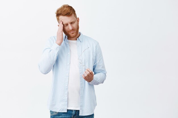 Retrato de hombre de negocios pelirrojo sombrío agotado con camisa azul casual, frotándose la sien, quitándose las gafas, trabajando en exceso, sufriendo dolor de cabeza sobre la pared gris