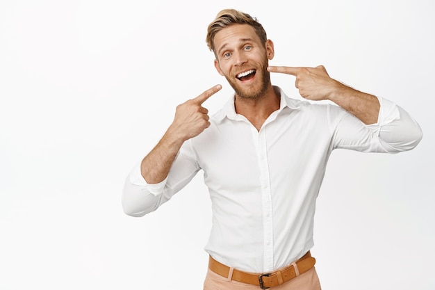 Retrato de un hombre de negocios señalando sus dientes blancos con una sonrisa blanqueada perfecta, un anuncio de una clínica dental de pie sobre un fondo blanco.