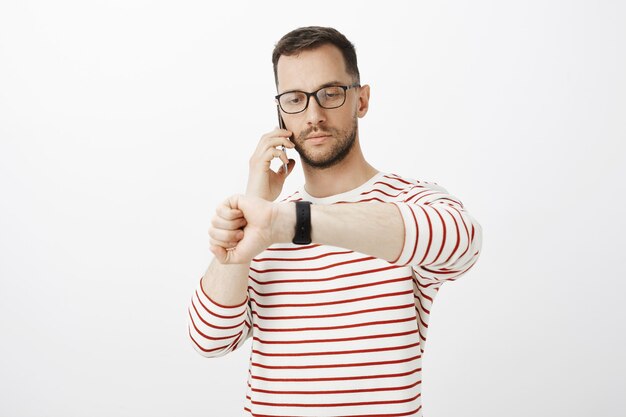 Retrato de hombre de negocios serio y guapo con gafas, hablando por teléfono inteligente y mirando el reloj, tratando con un socio comercial y decidiendo la hora de la reunión