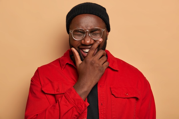 Retrato de hombre negro guapo cubre la mitad de la cara, se ríe positivamente, bromea con amigos y se divierte, usa sombrero negro y camisa roja