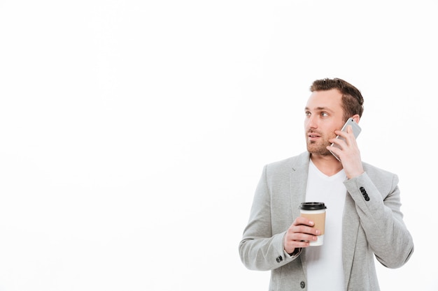 Retrato de hombre oficinista bebiendo café para llevar mientras tiene una agradable conversación móvil por teléfono celular y mirando a un lado, sobre la pared blanca