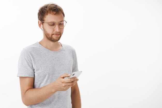 Retrato de hombre ordinario despreocupado relajado con cerdas en gafas navegando en el teléfono inteligente con auriculares inalámbricos recogiendo canciones mirando en la pantalla del dispositivo
