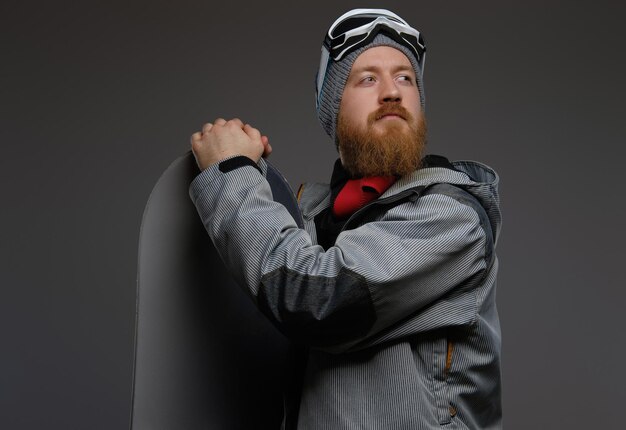 Retrato de un hombre pelirrojo con barba que lleva un equipo completo sosteniendo una tabla de snowboard y mirando hacia otro lado, aislado en un fondo oscuro.