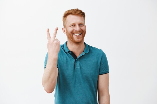 Retrato de hombre pelirrojo feliz y alegre de aspecto amable con barba haciendo el signo dos veces y sonriendo ampliamente mientras está de pie sobre la pared gris