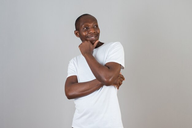 Retrato de hombre de piel oscura con camiseta blanca de pie con los brazos cruzados sobre la pared gris