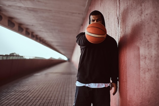 El retrato de un hombre de piel oscura vestido con una capucha negra y pantalones cortos deportivos sostiene una pelota de baloncesto mientras está parado en una acera debajo de un puente.