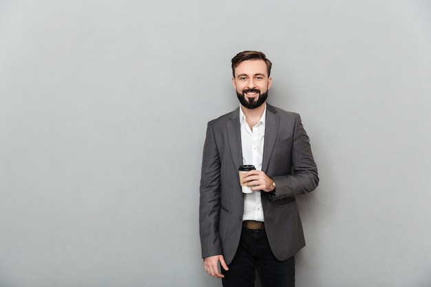 Retrato de hombre positivo en camisa blanca posando en la cámara con una amplia sonrisa y sosteniendo café para llevar sobre gris