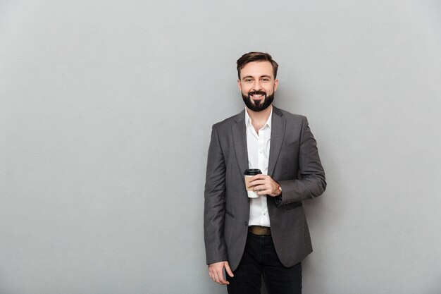 Retrato de hombre positivo en camisa blanca posando en la cámara con una amplia sonrisa y sosteniendo café para llevar sobre gris