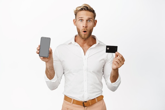 Retrato de un hombre que sostiene una tarjeta de crédito y muestra la pantalla de un teléfono móvil haciendo una expresión de cara sorprendida WOW mirando de pie sobre fondo blanco