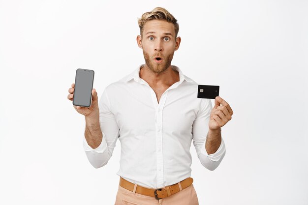 Retrato de un hombre que sostiene una tarjeta de crédito y muestra la pantalla de un teléfono móvil haciendo una expresión de cara sorprendida WOW mirando de pie sobre fondo blanco