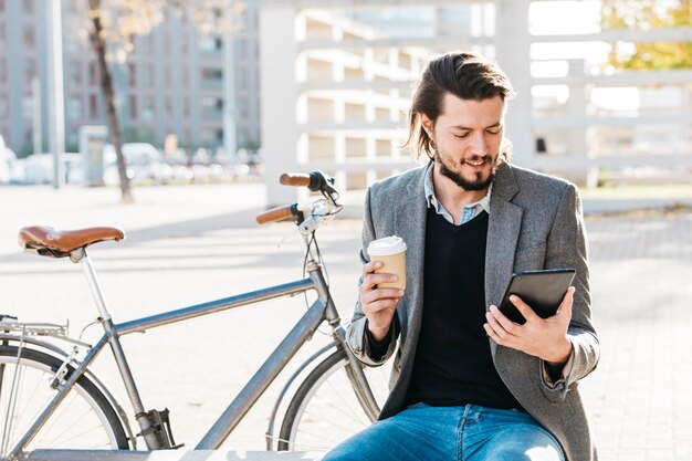 Retrato de un hombre que sostiene la taza de café disponible que mira el teléfono elegante que se sienta cerca de la bicicleta