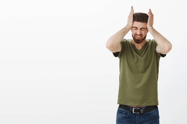 Retrato de hombre que sufre un gran dolor de cabeza o migraña agarrando la cabeza con ambas manos entrecerrando los ojos por el dolor y la angustia estando molesto y estresado de pie sobre una pared blanca infeliz