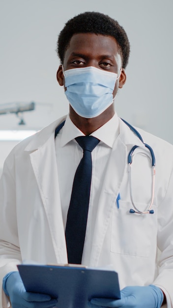 Retrato de un hombre que trabaja como médico con estetoscopio y archivos, de pie con mascarilla en la sala del hospital. Médico general con bata blanca y documentos de control durante la pandemia.