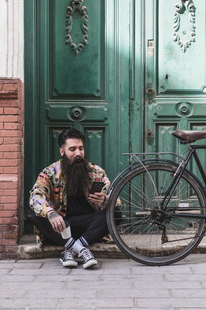 Retrato de un hombre que usa el teléfono móvil sentado cerca de la bicicleta frente a la puerta de madera verde