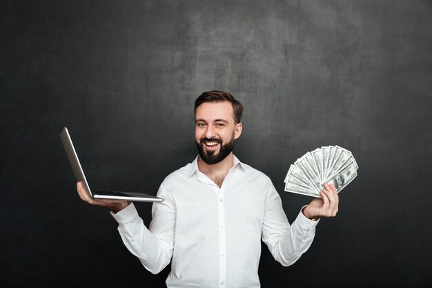 Retrato de hombre rico alegre en camisa blanca ganando mucho dinero en dólares con su cuaderno sobre gris oscuro