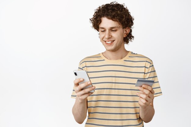 Retrato de hombre rizado sonriente compra en línea mirando el teléfono móvil con tarjeta de crédito de pie sobre fondo blanco.