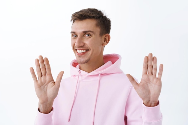 Foto gratuita retrato de hombre rubio sonriente guapo con sudadera con capucha rosa, levantando las manos en señal de rendición o gesto de disculpa, rechazando o rechazando la oferta cortésmente, de pie en la pared blanca